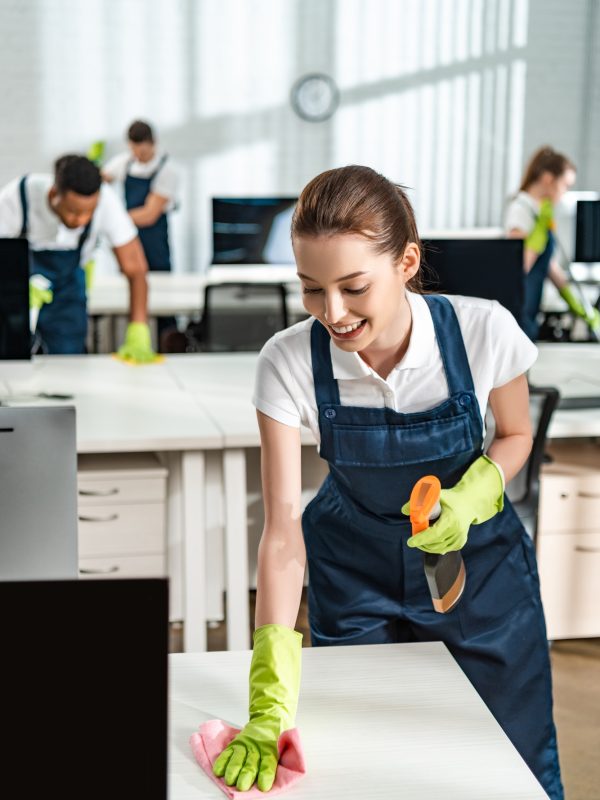 cheerful cleaner in overalls cleaning office desk with rag